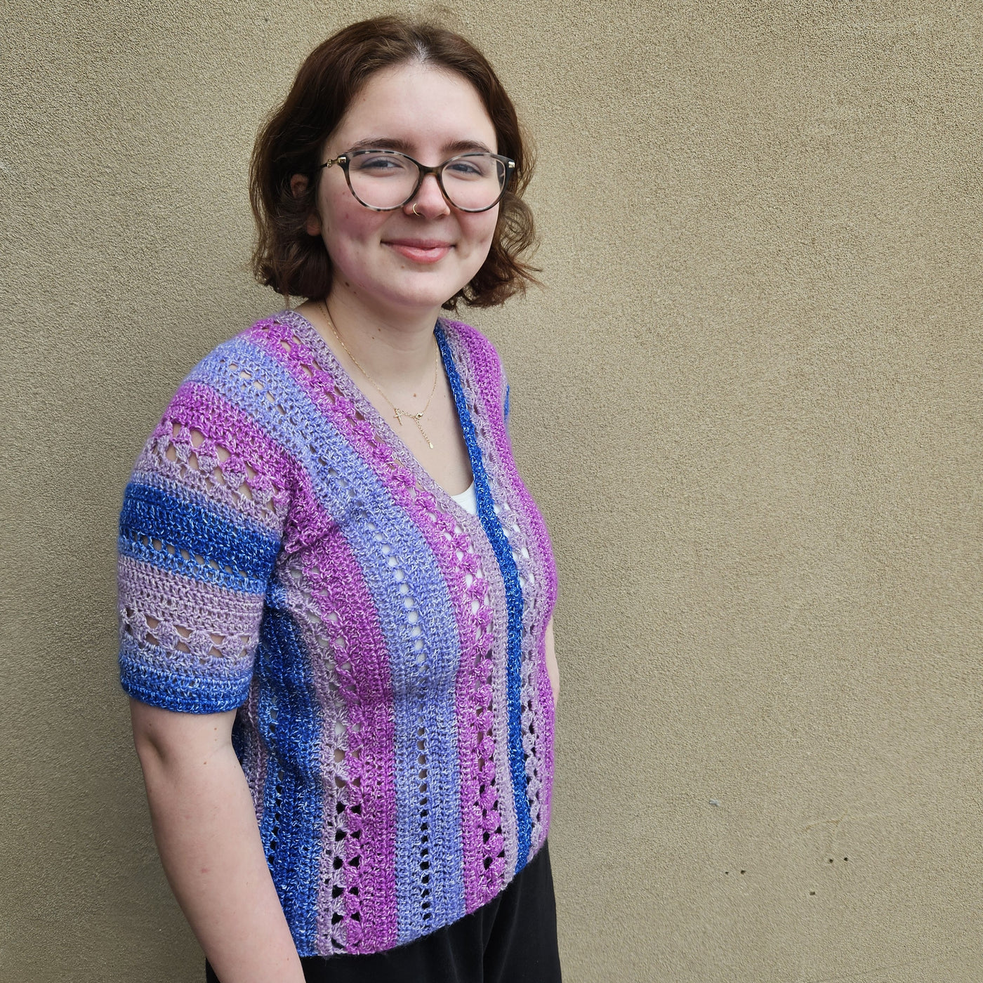 A woman wearing a colorful crocheted short-sleeve top stands against a textured beige wall. The top features vertical stripes in pink, purple, blue, and white with openwork crochet details.