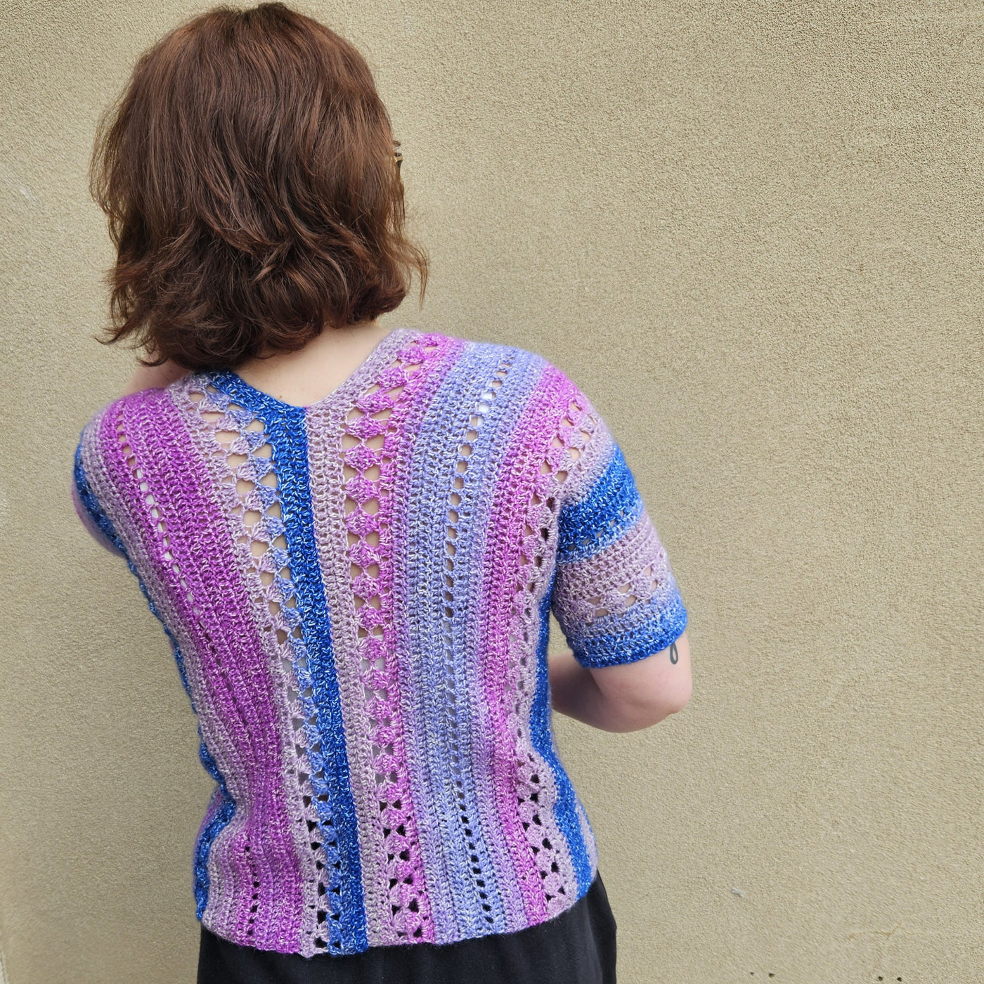 A woman wearing a colorful crocheted short-sleeve top stands against a textured beige wall. The top features vertical stripes in pink, purple, blue, and white with openwork crochet details.