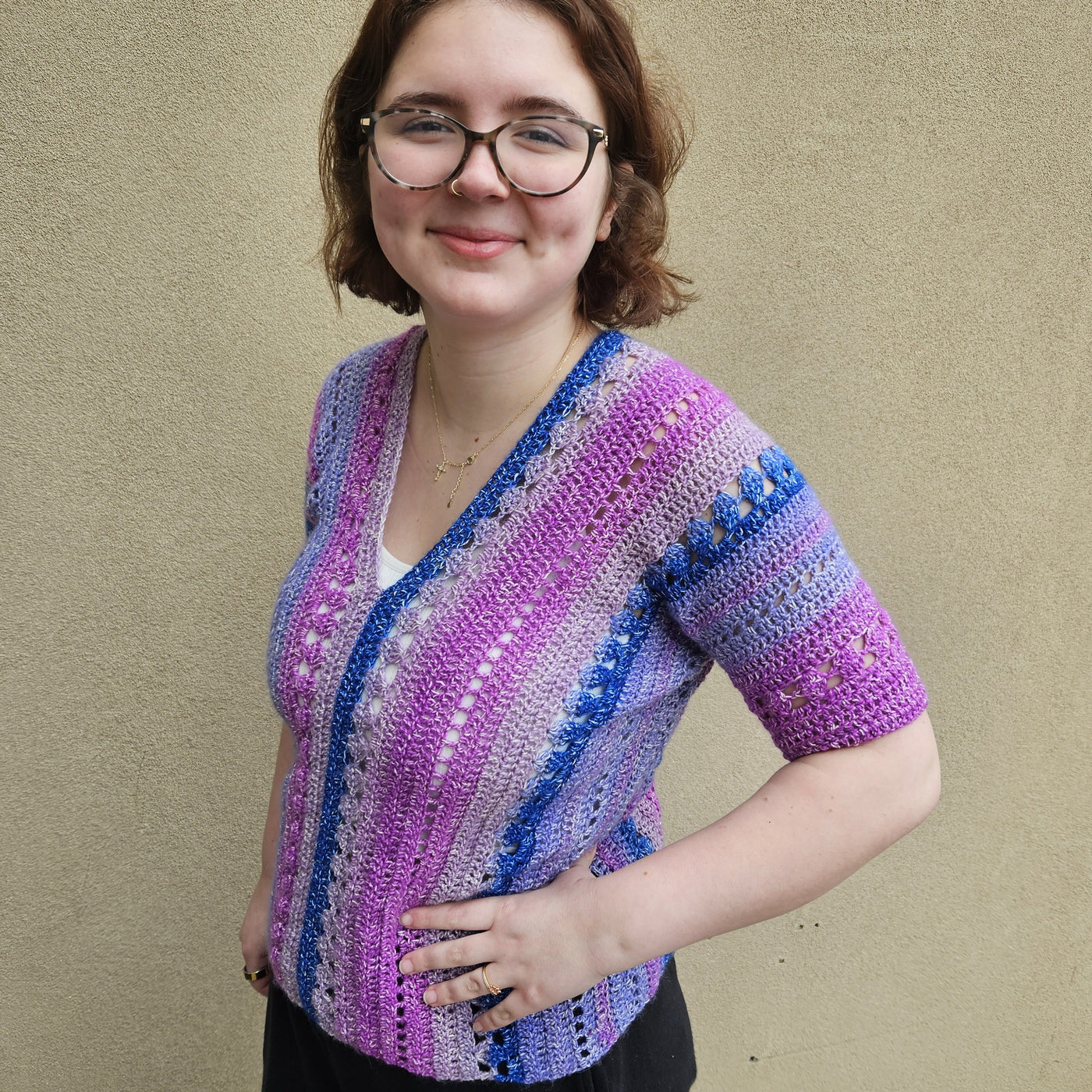 A woman wearing a colorful crocheted short-sleeve top stands against a textured beige wall. The top features vertical stripes in pink, purple, blue, and white with openwork crochet details.