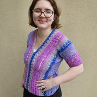 A woman wearing a colorful crocheted short-sleeve top stands against a textured beige wall. The top features vertical stripes in pink, purple, blue, and white with openwork crochet details.