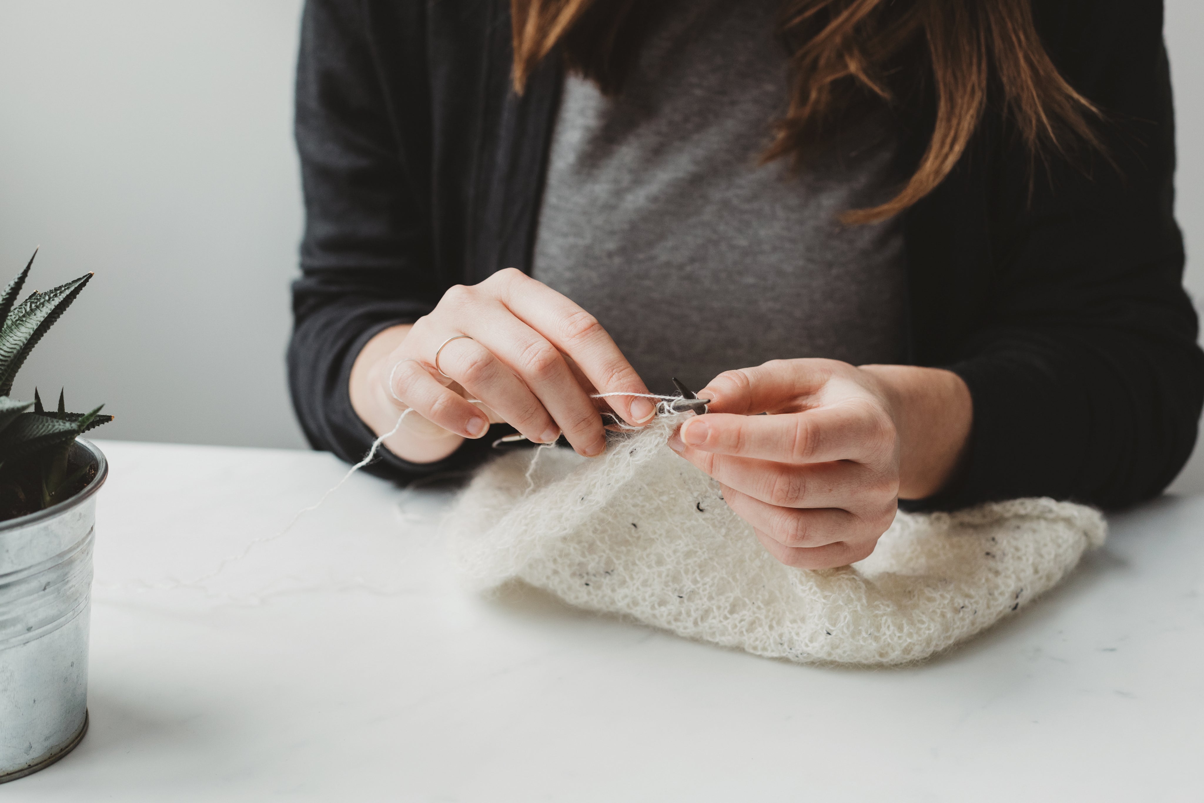 A woman knitting a white blanket on a white table with a plant nearby